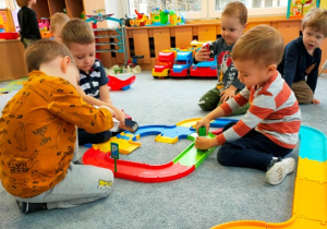 Chłopcy bawią się resorakami wykorzystując w zabawie plastikowy tor samochodowy ułożony na dywanie.